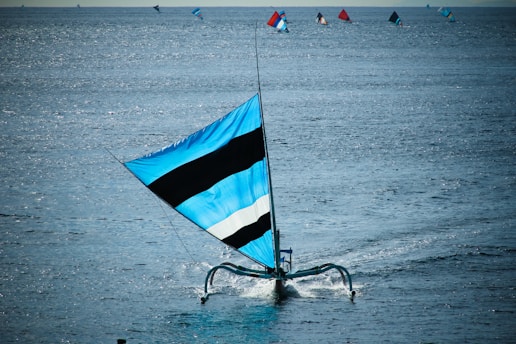 A sailboat with a large blue and black striped sail glides across a vast expanse of calm water. Several other sailboats with colorful sails are visible in the background, scattered across the sea. Sunlight reflects off the water, creating a shimmering effect.