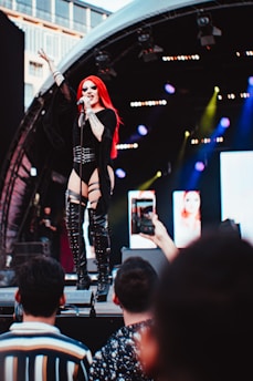 A performer with long red hair and black clothing sings passionately on stage. They are holding a microphone and are in front of a backdrop of lights and screens. The stage is partially covered by an arch, and audience members can be seen taking photos with their phones.