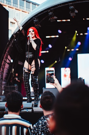 A performer with long red hair and black clothing sings passionately on stage. They are holding a microphone and are in front of a backdrop of lights and screens. The stage is partially covered by an arch, and audience members can be seen taking photos with their phones.