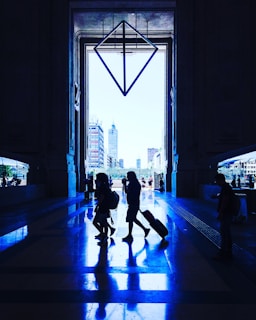 A large, grand entrance with towering doorways frames a cityscape view in the background. Silhouettes of people walking, some with luggage, create reflections on the glossy floor, highlighting the symmetry of the architecture. A geometric, diamond-shaped structure hangs prominently in the center, adding a modern artistic touch to the spacious setting.