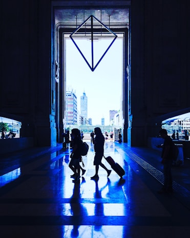 A large, grand entrance with towering doorways frames a cityscape view in the background. Silhouettes of people walking, some with luggage, create reflections on the glossy floor, highlighting the symmetry of the architecture. A geometric, diamond-shaped structure hangs prominently in the center, adding a modern artistic touch to the spacious setting.