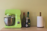 A vibrant green kitchen mixer is placed on a light wooden surface, accompanied by three bottles of varying colors and sizes. Next to the bottles, there is a paper towel holder with a roll of white paper towels.