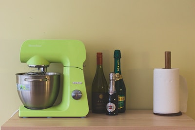 A vibrant green kitchen mixer is placed on a light wooden surface, accompanied by three bottles of varying colors and sizes. Next to the bottles, there is a paper towel holder with a roll of white paper towels.
