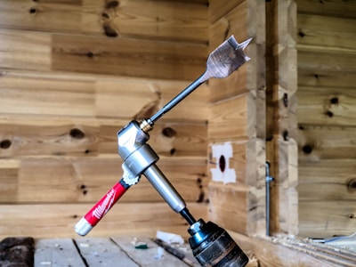 A spade drill bit is attached to a right-angle drill adapter, which is connected to a hand drill. The setup is placed against a wooden wall with a natural finish, showcasing visible grain patterns. The background includes woodwork details, adding a workshop or construction site atmosphere.