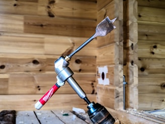 A spade drill bit is attached to a right-angle drill adapter, which is connected to a hand drill. The setup is placed against a wooden wall with a natural finish, showcasing visible grain patterns. The background includes woodwork details, adding a workshop or construction site atmosphere.