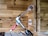 A spade drill bit is attached to a right-angle drill adapter, which is connected to a hand drill. The setup is placed against a wooden wall with a natural finish, showcasing visible grain patterns. The background includes woodwork details, adding a workshop or construction site atmosphere.
