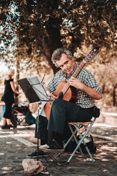 An older man sits on a folding chair, playing a classical guitar. He is focused on the sheet music placed on a stand in front of him. He is outdoors under a large tree, providing ample shade. The ground is cobblestoned, and a woman walks by in the blurred background.