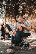 An older man sits on a folding chair, playing a classical guitar. He is focused on the sheet music placed on a stand in front of him. He is outdoors under a large tree, providing ample shade. The ground is cobblestoned, and a woman walks by in the blurred background.