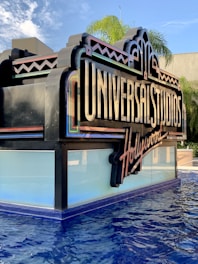 A large, decorative sign with 'Universal Studios Hollywood' written on it, positioned near a pool of water. The sign features bright and colorful elements with an art deco style. In the background, palm trees and a blue sky with some clouds add to the scenery.