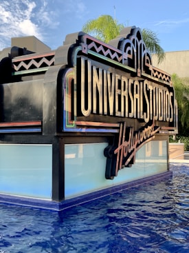 A large, decorative sign with 'Universal Studios Hollywood' written on it, positioned near a pool of water. The sign features bright and colorful elements with an art deco style. In the background, palm trees and a blue sky with some clouds add to the scenery.