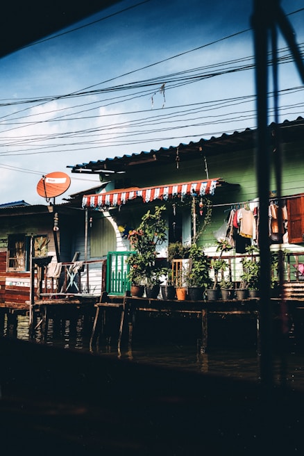 A rustic riverside scene featuring a wooden house with exterior walls painted in shades of green and white. The roof is covered with corrugated metal sheets, and a series of potted plants line the railing of a small porch. Above the house, a tangle of electrical wires crisscross the sky. Brightly colored clothes hang drying on lines, and a red satellite dish is attached to the side of the building.