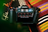 A vintage Nishika 3D N8000 camera is placed on a colorful woven textile background. The camera, with its distinctive quad-lens system, is prominently displayed with a strap attached.