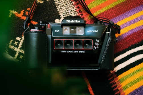 A vintage Nishika 3D N8000 camera is placed on a colorful woven textile background. The camera, with its distinctive quad-lens system, is prominently displayed with a strap attached.