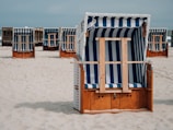 A beach scene with multiple wooden beach chairs covered by blue and white striped canopies. The chairs are arranged on pale sandy ground under a clear sky, with some chairs in the foreground and others in the background.