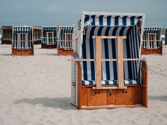 A beach scene with multiple wooden beach chairs covered by blue and white striped canopies. The chairs are arranged on pale sandy ground under a clear sky, with some chairs in the foreground and others in the background.