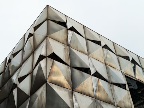 A building facade featuring a geometric pattern made of triangular panels, creating an angular, three-dimensional effect. The surface of the panels appears metallic, with varying shades of gray and hints of gold, reflecting light and creating a dynamic visual texture.