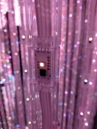 A close-up view of an LED strip light, attached to a transparent support, set against a background of numerous similar columns wrapped with glowing lights. The LED displays a soft pink hue, contributing to the overall ethereal ambiance.