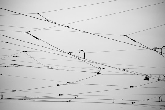 Numerous overhead electrical wires crisscross against a gray sky, creating an intricate and geometric pattern. The wires vary in thickness and tension, some having connectors and insulators attached.