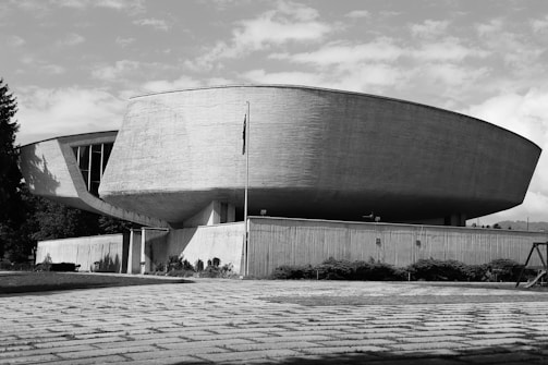 A large, futuristic building with a distinct, curved concrete structure. The architecture is modernist, with smooth lines and a sleek design. The setting appears to be outdoors with a paved area in the foreground and some shrubbery near the base of the building.