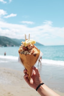 A hand holding a paper cone filled with seafood, garnished with a lemon slice. The background features a sandy beach with people in the distance and a calm blue sea under a clear sky.