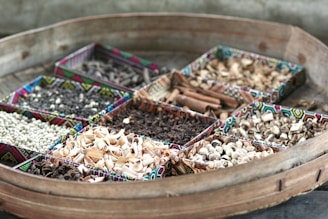 An assortment of spices is organized in a circular wooden tray. The spices are divided into separate sections using decorative, colorful fabric dividers, showcasing a variety of textures and colors.