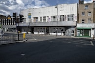A deserted urban street scene featuring a large, old building with graffiti on the shutters spelling 'HOMELESSNESS?'. The facade bears signs of disrepair and neglect. Nearby shops include a Chinese medicine store. A few people walk by, one with a dog, and traffic lights and street signs are visible.