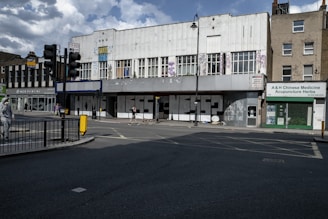 A deserted urban street scene featuring a large, old building with graffiti on the shutters spelling 'HOMELESSNESS?'. The facade bears signs of disrepair and neglect. Nearby shops include a Chinese medicine store. A few people walk by, one with a dog, and traffic lights and street signs are visible.