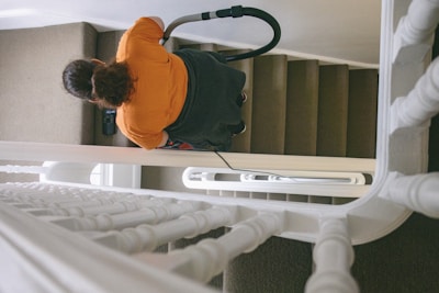 A person with dark hair wearing an orange shirt is vacuuming a carpeted staircase. The view is from above, showcasing the banister and several steps. The vacuum cleaner's hose and handle are visible as the person cleans the stairs.