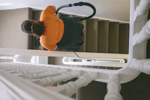 A person with dark hair wearing an orange shirt is vacuuming a carpeted staircase. The view is from above, showcasing the banister and several steps. The vacuum cleaner's hose and handle are visible as the person cleans the stairs.