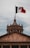 A monumental building featuring classical architecture with stone columns and an ornate domed roof. Above the building, a large Mexican flag with red, white, and green stripes is hoisted on a tall flagpole, prominently displaying its national emblem.