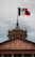 A monumental building featuring classical architecture with stone columns and an ornate domed roof. Above the building, a large Mexican flag with red, white, and green stripes is hoisted on a tall flagpole, prominently displaying its national emblem.