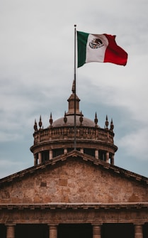 A monumental building featuring classical architecture with stone columns and an ornate domed roof. Above the building, a large Mexican flag with red, white, and green stripes is hoisted on a tall flagpole, prominently displaying its national emblem.