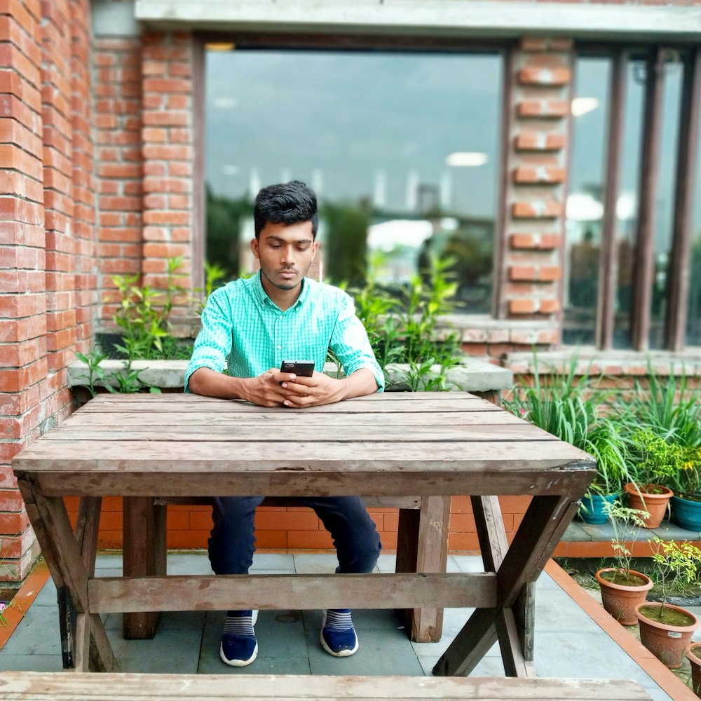 man wearing green collared button-up long-sleeved shirt using phone while sitting near brown wooden table