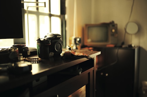 A dimly lit room features a digital camera resting on a wooden desk, surrounded by various objects such as camera lenses and small trinkets. A vintage television set and other electronics are visible in the background, with sunlight streaming through a nearby window, creating an atmosphere of nostalgia and creativity.