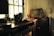 A dimly lit room features a digital camera resting on a wooden desk, surrounded by various objects such as camera lenses and small trinkets. A vintage television set and other electronics are visible in the background, with sunlight streaming through a nearby window, creating an atmosphere of nostalgia and creativity.