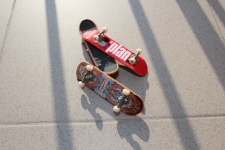 Two miniature skateboards are placed on a speckled, light-colored floor. One skateboard has a bright red top with a bold white design, while the other features an intricate, colorful pattern. The image is well-lit, casting defined shadows of the skateboards and their components onto the floor.