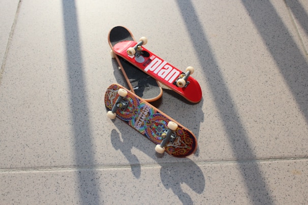 Two miniature skateboards are placed on a speckled, light-colored floor. One skateboard has a bright red top with a bold white design, while the other features an intricate, colorful pattern. The image is well-lit, casting defined shadows of the skateboards and their components onto the floor.
