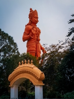 A large, orange-colored statue of a Hindu deity stands prominently amidst greenery. The statue has its right hand raised in a gesture of blessing and its left hand resting on the waist, adorned with traditional attire and ornaments. Below the statue is an arched entryway that is similarly styled in an orange color, supported by two white pillars. Trees and foliage surround both the statue and the entryway, creating a natural backdrop.