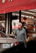 An older man stands in front of a store with a red awning, appearing thoughtful. People are visible entering or exiting the store, which has products displayed on tables with colorful signs. A car is parked close by, with a California license plate visible.
