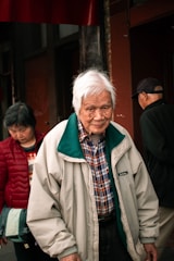 An elderly man with white hair wearing a light-colored jacket and checkered shirt stands outdoors with two other individuals. The background consists of a building with a reddish hue.