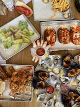 A variety of seafood dishes are artfully arranged on metal trays and plates over a wooden table. The spread includes fresh oysters on ice garnished with lemon wedges and dipping sauces, lobster rolls, crispy fried seafood, shrimp cocktail, and a Caesar salad with anchovies. Sea urchins with their distinctive spiky shells are also present, opened to reveal their contents. French fries appear in a tray accompanied by sauces. The setting suggests a casual and abundant seafood meal.