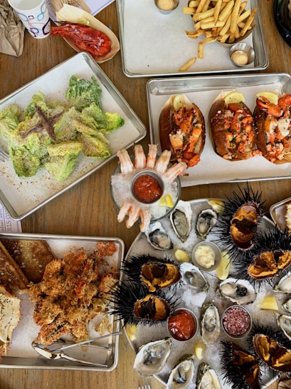 A variety of seafood dishes are artfully arranged on metal trays and plates over a wooden table. The spread includes fresh oysters on ice garnished with lemon wedges and dipping sauces, lobster rolls, crispy fried seafood, shrimp cocktail, and a Caesar salad with anchovies. Sea urchins with their distinctive spiky shells are also present, opened to reveal their contents. French fries appear in a tray accompanied by sauces. The setting suggests a casual and abundant seafood meal.