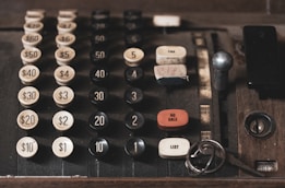 An old, vintage cash register with large, mechanical buttons displaying different denominations of money from $1 to $60. The cash register features buttons for functions like 'NO SALE', 'TAX', and 'LIST'. There are keys resting near the lower right corner of the cash register.