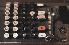 An old, vintage cash register with large, mechanical buttons displaying different denominations of money from $1 to $60. The cash register features buttons for functions like 'NO SALE', 'TAX', and 'LIST'. There are keys resting near the lower right corner of the cash register.