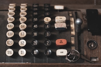 An old, vintage cash register with large, mechanical buttons displaying different denominations of money from $1 to $60. The cash register features buttons for functions like 'NO SALE', 'TAX', and 'LIST'. There are keys resting near the lower right corner of the cash register.