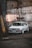 A vintage, white Volkswagen GTI is parked in an industrial setting against the backdrop of a weathered concrete wall with visible stains and cracks. The car features distinctive round headlights and custom white rims that give it a classic look. The ambiance is accented by dim industrial lighting and a rugged surface.