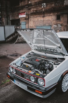 A white Volkswagen GTI with its hood open reveals an Audi engine. The car is parked in an industrial setting with a weathered, urban background that includes aged walls and a metal gate.