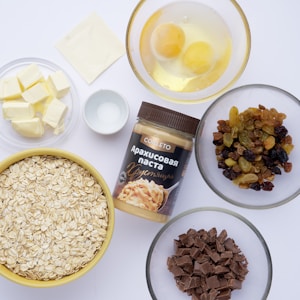 A collection of baking ingredients is arranged on a white surface, including a jar of peanut butter, oats in a yellow bowl, chopped chocolate, dried mixed fruits, cubes of butter, two eggs in a bowl, and a small dish of salt. A square of parchment paper is also visible.