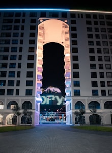 A modern multi-story building with an arched structure in the center. The building's exterior is illuminated with various lights. A smaller structure with LED lights displaying 'DFW' can be seen through the arch.