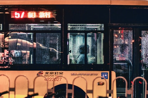 A passenger sits inside a bus on a rainy night. The bus windows are covered with raindrops, reflecting colorful city lights. The interior is dimly lit, highlighting the solitary figure looking out. A digital display shows route information in red and yellow text.
